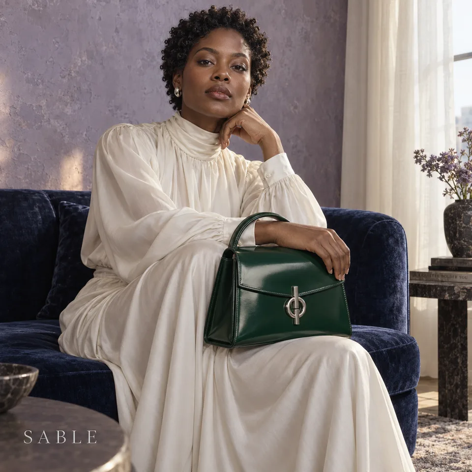Square luxury fashion photo of an original adult Black woman with short natural curls seated upright on a low indigo velvet sofa, looking directly at camera. Fully covered flowing ivory silk dress with high neckline and long sleeves