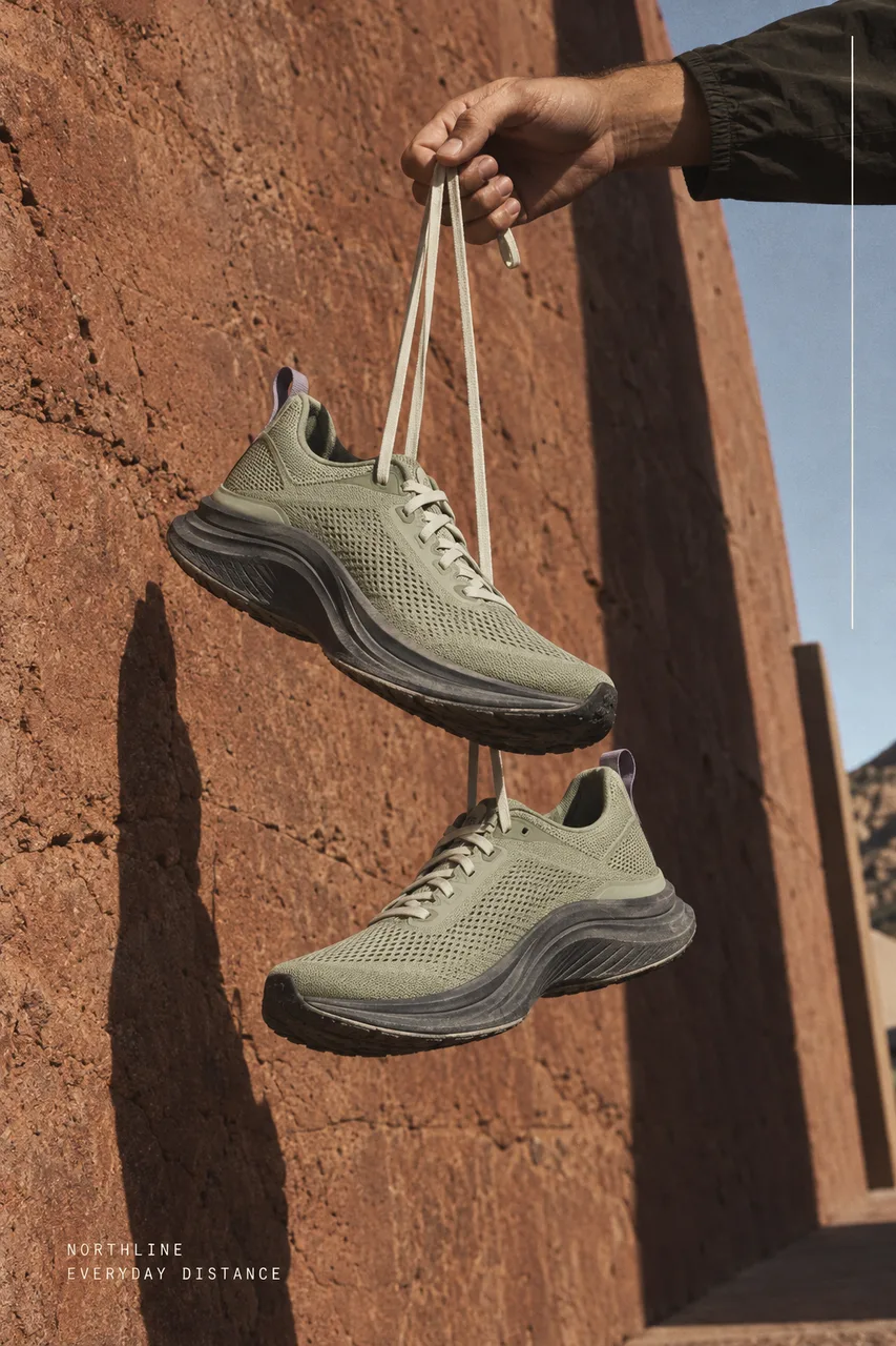 Portrait running campaign, photographed from ground level. Two unbranded pale-olive mesh running shoes hang from white laces held by an adult hand entering from upper-right, against rough rust-red sandstone and a long narrow architectural shadow