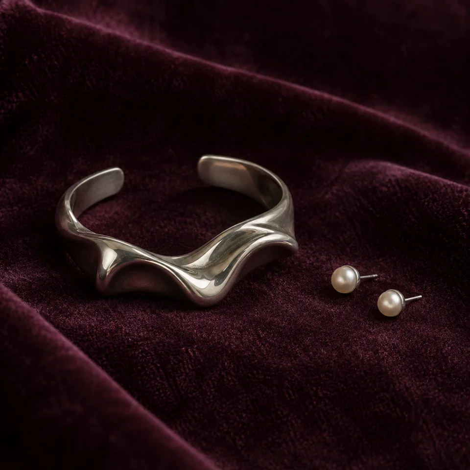 Square close product photo of an original sculptural silver cuff and tiny pearl earrings on deep plum velvet, restrained side lighting, soft shadow and realistic metal reflections. Tactile velvet pile, elegant negative space, no logos or text.